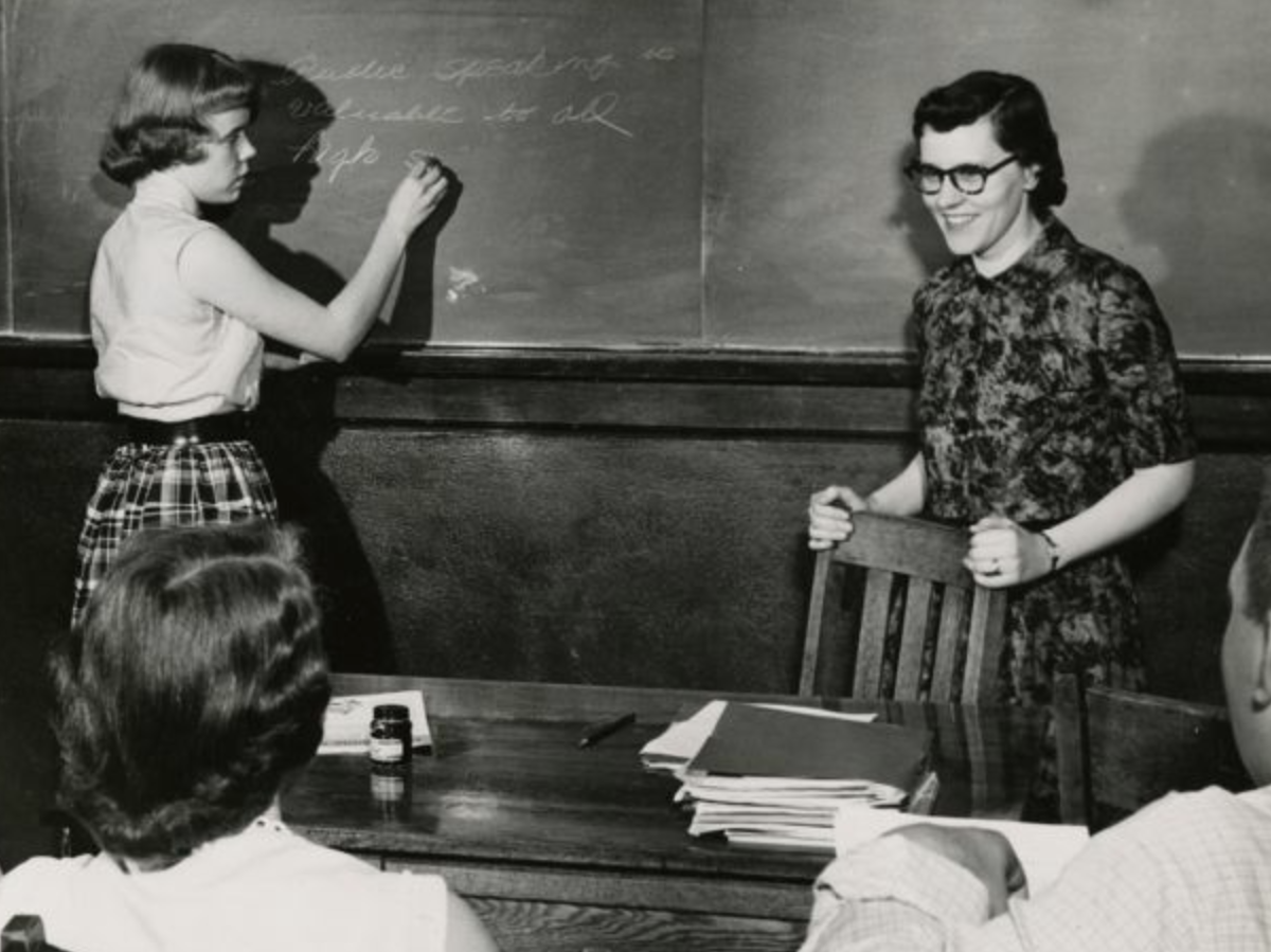 Female student writing on chalkboard with smiling teacher looking at class, St. Paul, Minnesota