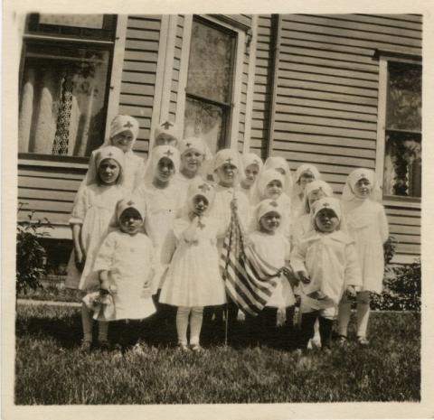 Children dressed as Red Cross nurses pose with an American flag