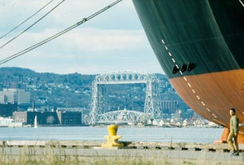 Tip of black-red ship and person walking. Far behind is Duluth's lift bridge
