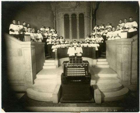 Around 40 boys on stone or wood stands, behind two men choir conductors who are behind a piano. All wearing white