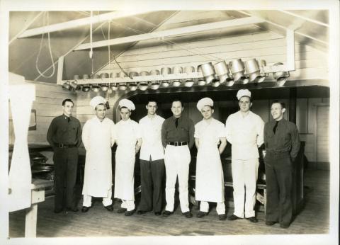Men in military style or white cooks uniforms stand in a kitchen under a line of pots and pans