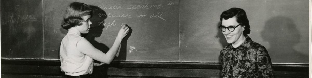 Female student writing on chalkboard with smiling teacher looking at class, St. Paul, c. 1940. Bethel University Archives.